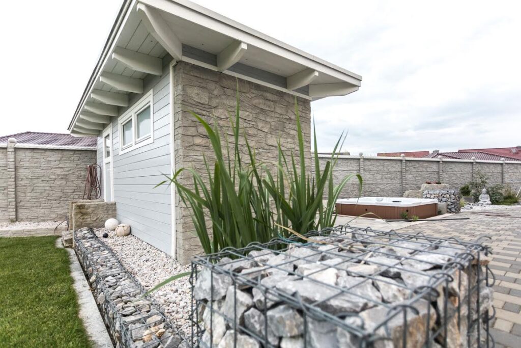 Moderner Garten mit Betonzaun und Nebengebäude. Steinoptik-Betonplatten umgeben den Bereich. Im Vordergrund ein Gabione mit Pflanzen, im Hintergrund ein Whirlpool. Helle Farbtöne und gepflegte Landschaftsgestaltung vermitteln einen eleganten Eindruck