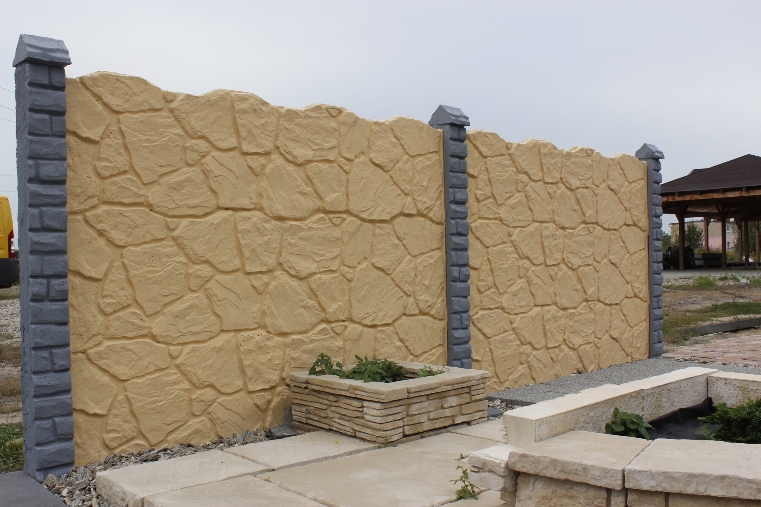 Ein dekorativer Betonzaun mit Felsoptik in sandfarbener Tönung. Die Wand besteht aus großen, unregelmäßigen Steinmustern und wird von grauen Betonsäulen gestützt. Im Vordergrund befindet sich ein kleines Steinbeet mit Pflanzen, umgeben von einer gepflasterten Fläche