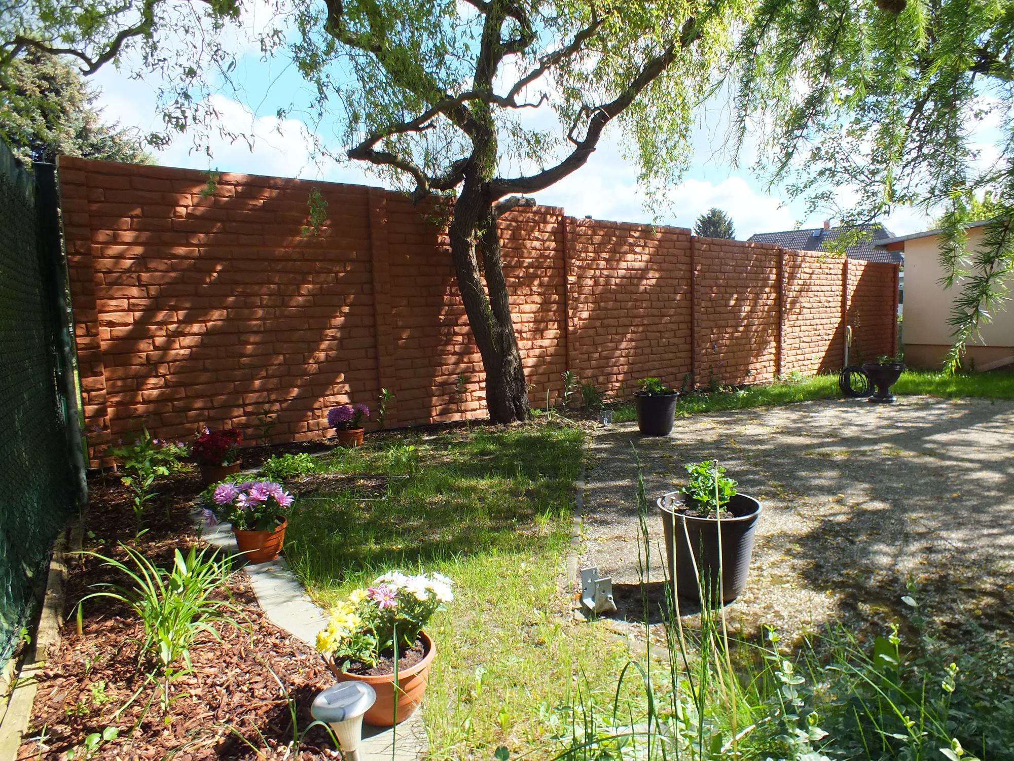 "Ein Garten mit einem langen Betonzaun in Ziegeloptik. Ein alter Baum wirft Schatten auf die Mauer. Im Vordergrund sieht man Blumentöpfe mit bunten Blumen und einen gepflegten Rasen. Die Szene vermittelt eine friedliche Gartenatmosphäre.