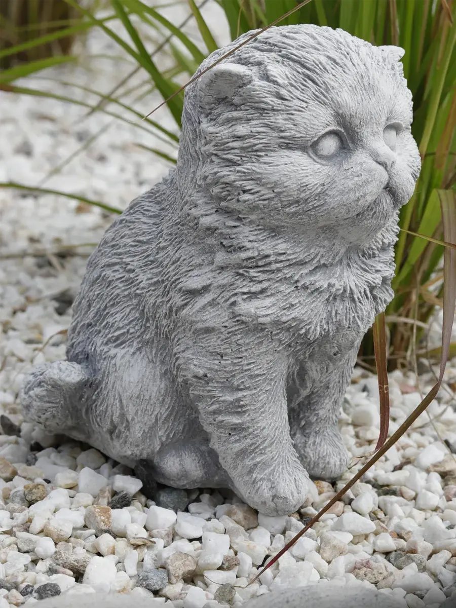 Graue Betonfigur einer flauschigen Katze im Garten, auf weißem Kies sitzend, umgeben von grünen Gräsern